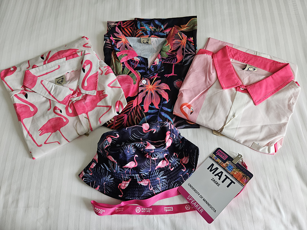 Collection of speaker attire, with one shirt for each day. All three contain a lot of pink and are emblazened with flamingos. A similarly themed hat and pink branded lanyard was included.