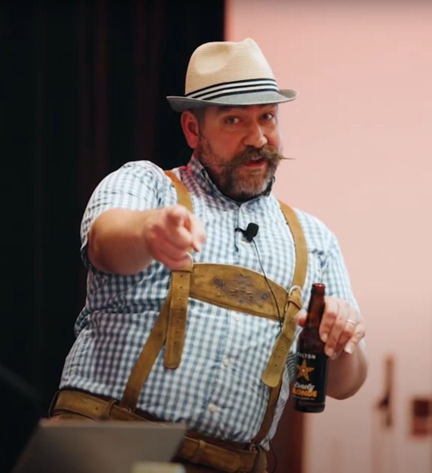 Screen snip of video footage taken during a Happy Hour session of Matt wearing lederhosen while on stage holding a beer.